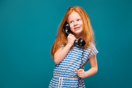 Isolated on blue, pretty, caucasian redhead child in blue striped dress, with long rusty hair hold black phone handset, curious, hold the wireの写真素材