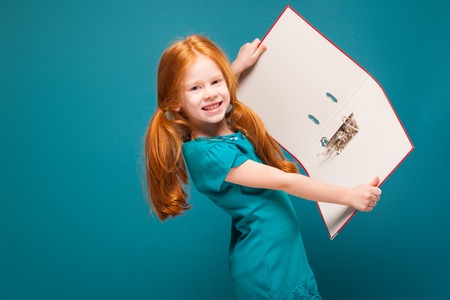 Isolated on blue, beautiful caucasian redhead child in azure dress, with long rusty hair, hold red paper folder, show folder to the cameraの写真素材