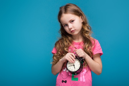 Isolated on blue, attractive caucasian brunette child with long hair, in pink t-shirt and blue jeans hold the alarm clock, shyの写真素材