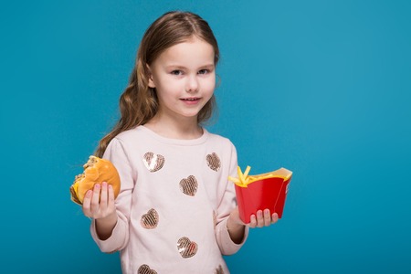 Isolated on blue, attractive caucasian brunette child with long hair, in beige coat and blue jeans hold hamburger and friesの写真素材