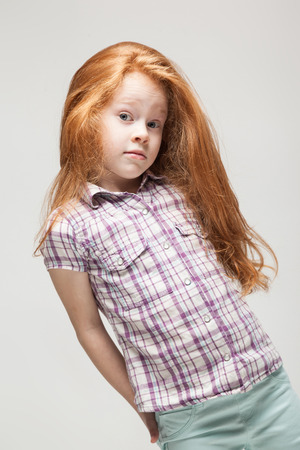 Vertical picture, isolated on grey, adorable, cute little caucasian redhead girl in plaid shirt, bright blue trousers and white boots, look at camera, turned, confused, hands on her backの写真素材