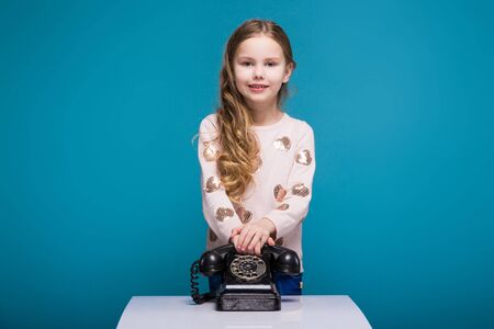 Isolated on blue, attractive caucasian brunette child with long hair, in beige coat and blue jeans hold black phone handset, look at cameraの写真素材