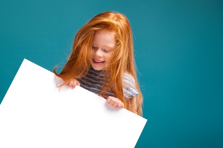 Isolated on blue, pretty, caucasian redhead child in blue striped dress, with long rusty hair show big empty poster, turned, look at posterの写真素材