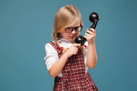 Isolated on blue, beautiful cute little caucasian blond girl in red dress, white shirt and glasses holds phone handset, look at handsetの写真素材