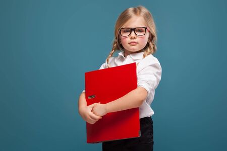 Isolated on blue, attractive cute little caucasian blond girl in white shirt, glasses and black trousers hold red paper folder, look at camera, unhappyの写真素材