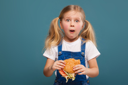 Isolated on blue, beautiful cute little caucasian blond girl in white shirt, white jacket, glasses and white shorts with hamburger, look at camera, amazedの写真素材