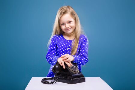 Isolated on blue, pretty little caucasian blond girl in purple dress holds black phoneの写真素材