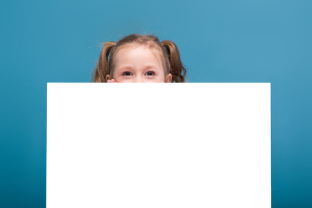 Isolated on blue, cute little caucasian brunette girl in pink shirt with monkey and blue trousers hold empty poster, hide behind poster, look at cameraの写真素材