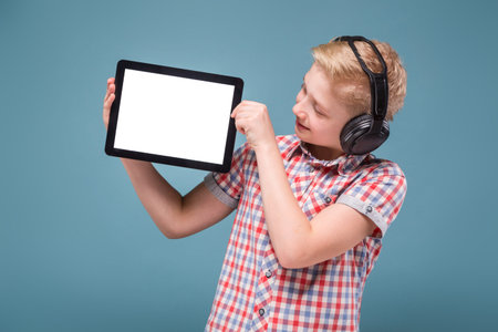 smiling blond student with headphones holding the phone at arms length and shows the display, the lap studio shot European teenager picture with depth of field, selective focus on tabletの写真素材