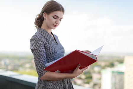 Blured sky and sity on background, attractive brunette caucasian businesslady in patterned dress and watch stand on the roof and hold red paper folder, look in folderの写真素材