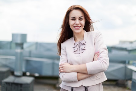 Blured sky on background, pretty redhead caucasian businesslady in beige suit stand on the roof, crossed hands, smilingの写真素材