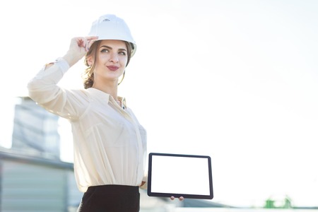 Blured roof on background, attractive brunette caucasian businesslady in white blouse, watch, white helmet and black skirt stand on the roof and show empty tablet, hand on helmetの写真素材