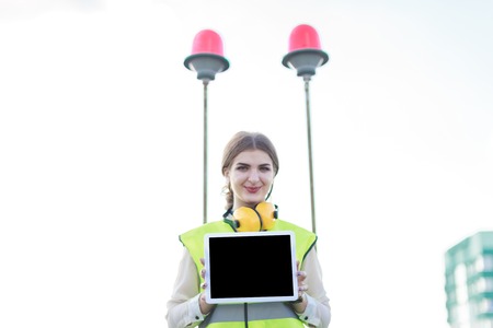 Blured sky and red signal lights on background, young pretty brunette caucasian woman in green west and yellow earmuffs stand on roof and show empty tablet in frontの写真素材