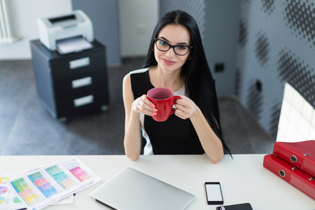 Office on background, beautiful young brunette caucasian businesswoman in black dress and glasses sit at the table and hold red cup, office stuff and laptop on the desk, from aboveの写真素材