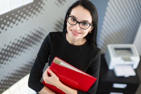 Office on background, beautiful young brunette caucasian businesswoman in black dress and glasses hold red paper folder, hugs folder, look at cameraの写真素材