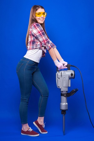 Isolated on blue, beautyful sexy brunette caucasian girl in chechered shirt, yellow build goggles, snickers and blue jeans stand near jackhammer, hold it with both hands, look at camera, smilingの写真素材