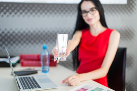 Office on background, pretty, young brunette caucasian businesslady in red dress and glasses sit at the table and work with laptop, water bottle on the desk, show glass, focus on glass, look at cameraの写真素材