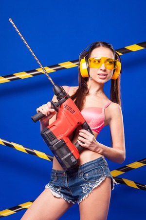 Isolated on blue, beauty brunette caucasian builder girl in denim shorts, white snickers, pink bikini and yellow build glasses stand with electro saw, look at leftの写真素材