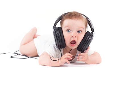 isolated on white, cute pretty caucasian baby boy, in white shirt lies on tummy wiith black headphones on head, listening music, amazedの写真素材