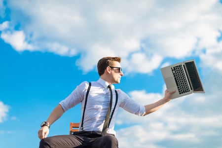 Attractive brunette caucasian businessman in white shirt, black tie, black braces, black trousers, costly watch and black sunglasses sit on a chair on the roof, going to drop his laptop, clouds on backgroundの写真素材