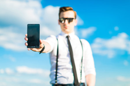 Serious attractive brunette caucasian businessman in white shirt, black tie, black braces, costly watch and black sunglasses stand on the green ladder on the roof and hold a cellphone, show phone to camera, look at camera, blue sky on backgroundの写真素材