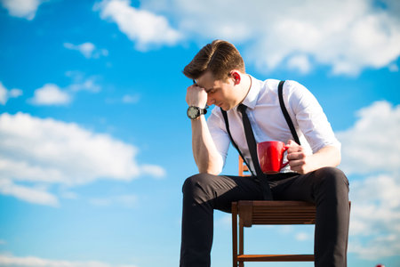 Attractive brunette caucasian businessman in white shirt, black tie, black braces, black trousers, costly watch and black sunglasses sit on a chair on the roof, hold red cup, hands on knees, in thoughtclouds on backgroundの写真素材