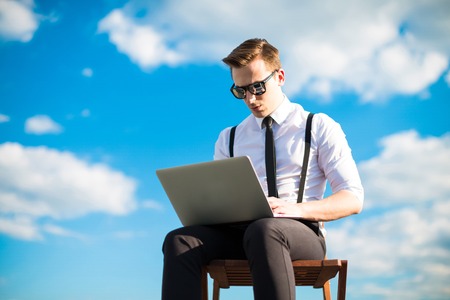 Attractive brunette caucasian businessman in white shirt, black tie, black braces, black trousers, costly watch and black sunglasses sit on a chair on the roof, work with black laptop, tapping
clouds on backgroundの写真素材