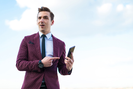 Attractive young brunette caucasian businessman in red business suit, white shirt, black tie, black trousers and costly watch stand on the roof and hold black cellphone, point at phoneblured city on backgroundの写真素材