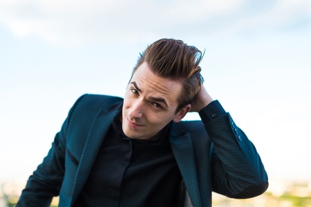 Young good looking brunette caucasian businessman in dark suit, black shirt and costly watch stand on the roof, look at camera, scratches his back of the head
blured city on backgroundの写真素材