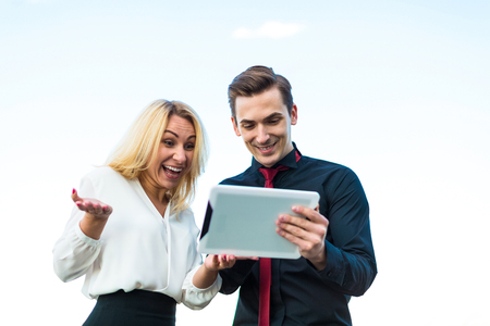 Couple of businessmen, young good looking brunette caucasian man in dark suit, red tie  and black shirt, and attractive blonde woman in white blouse and black skirt stand on the roof and hold one tablet, both look at tablet, joyfulblured city on backgroundの写真素材
