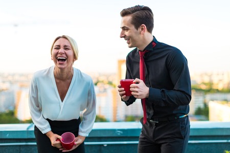 Couple of caucasian businessmen, young attractive brunette man in dark suit, red tie  and black shirt, and pretty blonde woman in white blouse and black skirt stand on the roof with red cups of coffe, having a conversation, woman is laughingblured city on backgroundの写真素材