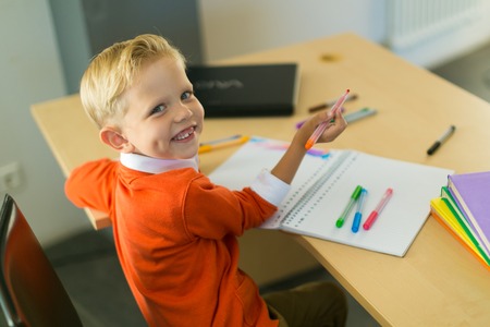 Cute blonde caucasian kid sit at the table with colorful folders and draws with pen, look at camera, hold red pencil, smiling, turnedの写真素材