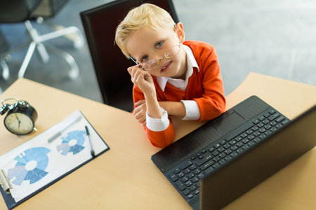 Young blonde caucasian kid in orange shirt sit at the table with colorful folders and alarm clock...