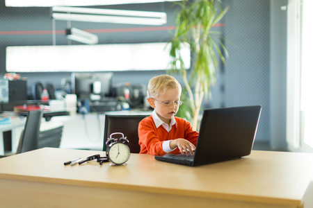 Young blonde caucasian kid in orange shirt sit at the table with colorful folders and alarm clock and plays on the black laptop
Blurred office on backgroundの写真素材