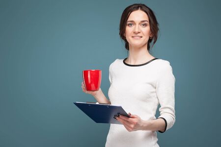 Isolated on dark grey, pretty young caucasian brunette woman dressed in white shirt stand and hold blue tablet and red cupの写真素材