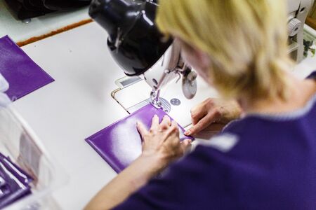blonde short-haired woman in dark-blue uniform stitching a purple wallet with a sewing machine. wallet is in focus.の写真素材