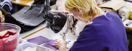 blonde short-haired woman in dark-blue uniform stitching a purple wallet with a sewing machine under...