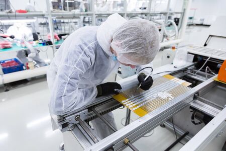 a factory worker in a white lab suit, black latex gloves and face mask, working with a soldering iron and some modern equipment.の写真素材