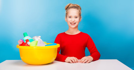 isolated on blue, smiling girl in red pullover with fair-haired updo, sitting at the white table with a lemon washbowl full of multicolored detergents and rags on it. copyspace.の写真素材