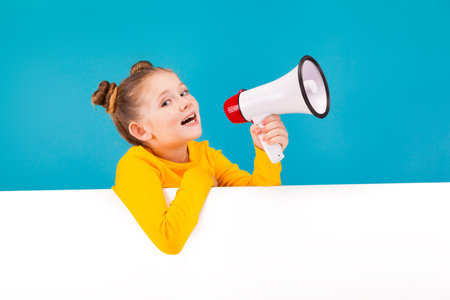 isolated on blue, adorable red-haired girl with freckles in lemon sweater with the white megaphone, looking into the camera. hands are over the white background. copyspace.の写真素材
