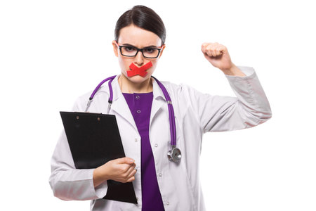 Portrait of young attractive female doctor in white coat standing in office looking in camera and holding black pad frustrated angry stressed sad disappointed with taped mouth as medical secret concept confident experienced skilful brunette caucasian beautiful pretty gp.の写真素材