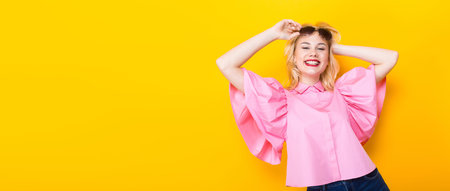 Portrait of cheerful laughing blonde woman with red lips in pink shirt and jeans shorts with sunglasses on orange background with copyspace touching her hair. Fashion advertisement concept horizontal.の写真素材