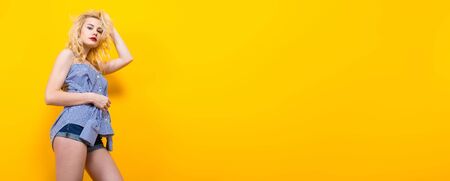 Portrait of attractive blonde woman with red lips in blue striped shirt and jeans shorts on yellow background with copyspace touching her hair. Fashion and beauty advertisement concept horizontal.の写真素材