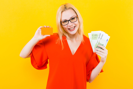 Portrait of woman in glasses and red dress isolated on yellow background hold blank credit card and fan of euro banknotes earn on the internet withdraw money loan mortgage bonus award win concept.の写真素材