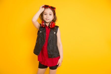 Portrait of little cute girl in red shirt, sunglasses holding earphones on her neck isolated on orange background .の写真素材