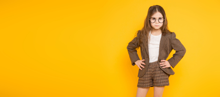 Portrait of adorable long-haired little girl in eyeglasses wearing brown jacket, shorts and white knee-socks with hands on waist isolated on orange background with copyspace kids fashion concept.の写真素材