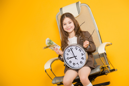Long-haired little girl in eyeglasses sits in chair wearing brown jacket, shorts and white knee-socks with watches and fan of euro bills isolated on orange background with copyspace time is money.の写真素材