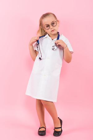 Portrait of little girl dressed like doctor in white coat and eyeglasses wearing stethoscope isolated on pink background with copyspace pediatrics concept.の写真素材