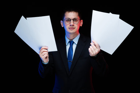 Young business man in black suit standing in office looking in camera with documents in his hands...