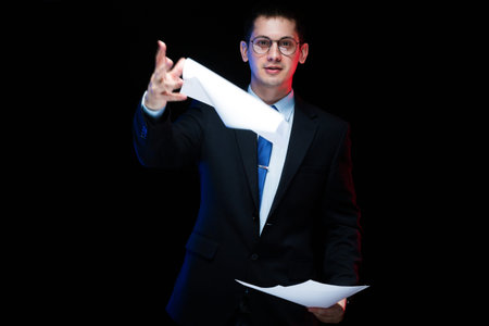 Young business man in black suit standing in office looking in camera with documents in his hands...
